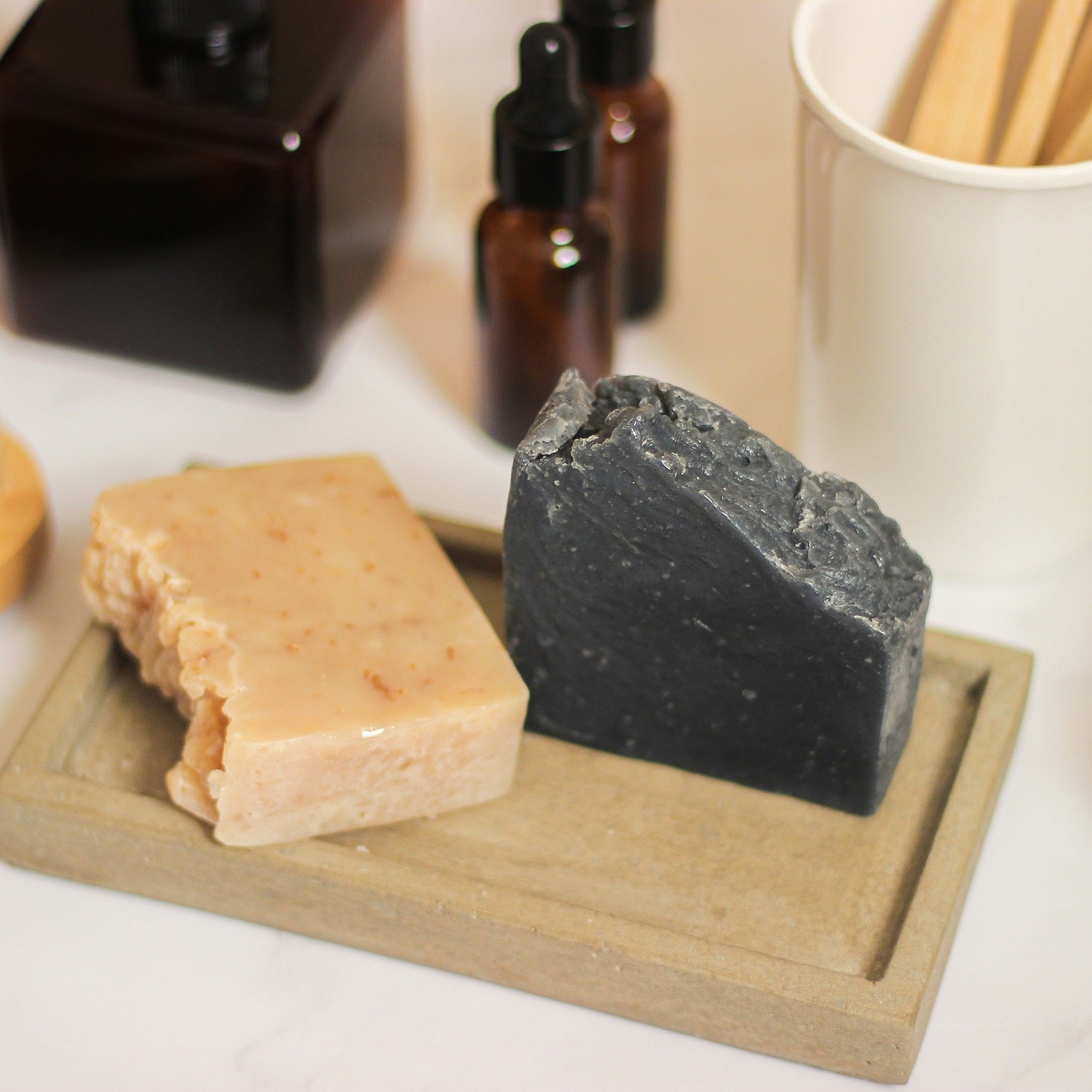 Two bars of soap, one beige and one black, on a wooden tray with blurred background.